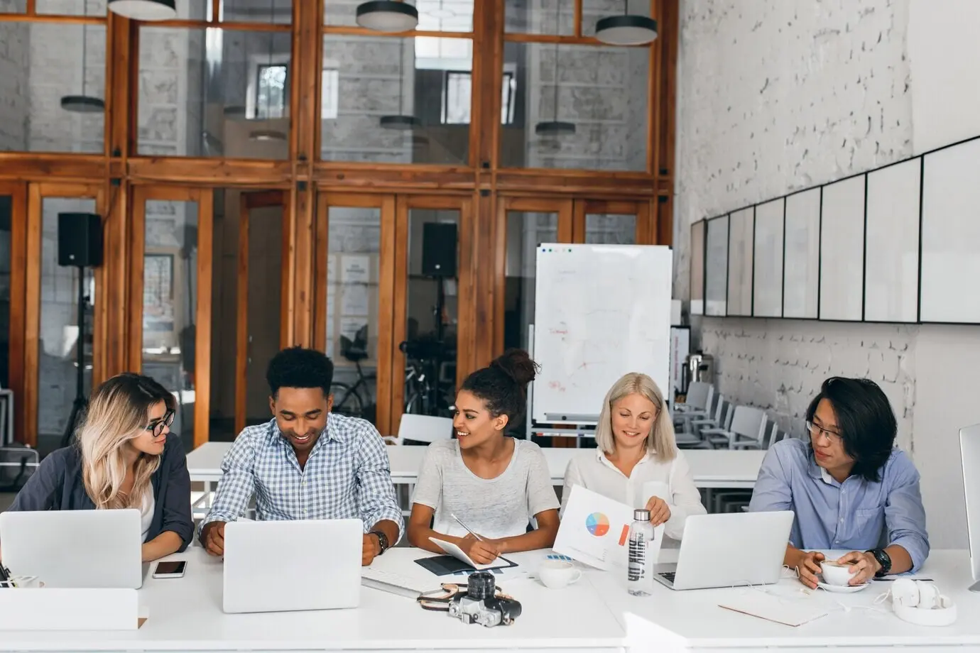 A tired Asian IT specialist drinks coffee and watches a female colleague work on a laptop. An indoor portrait of young business people sitting together at the table in a conference hall.