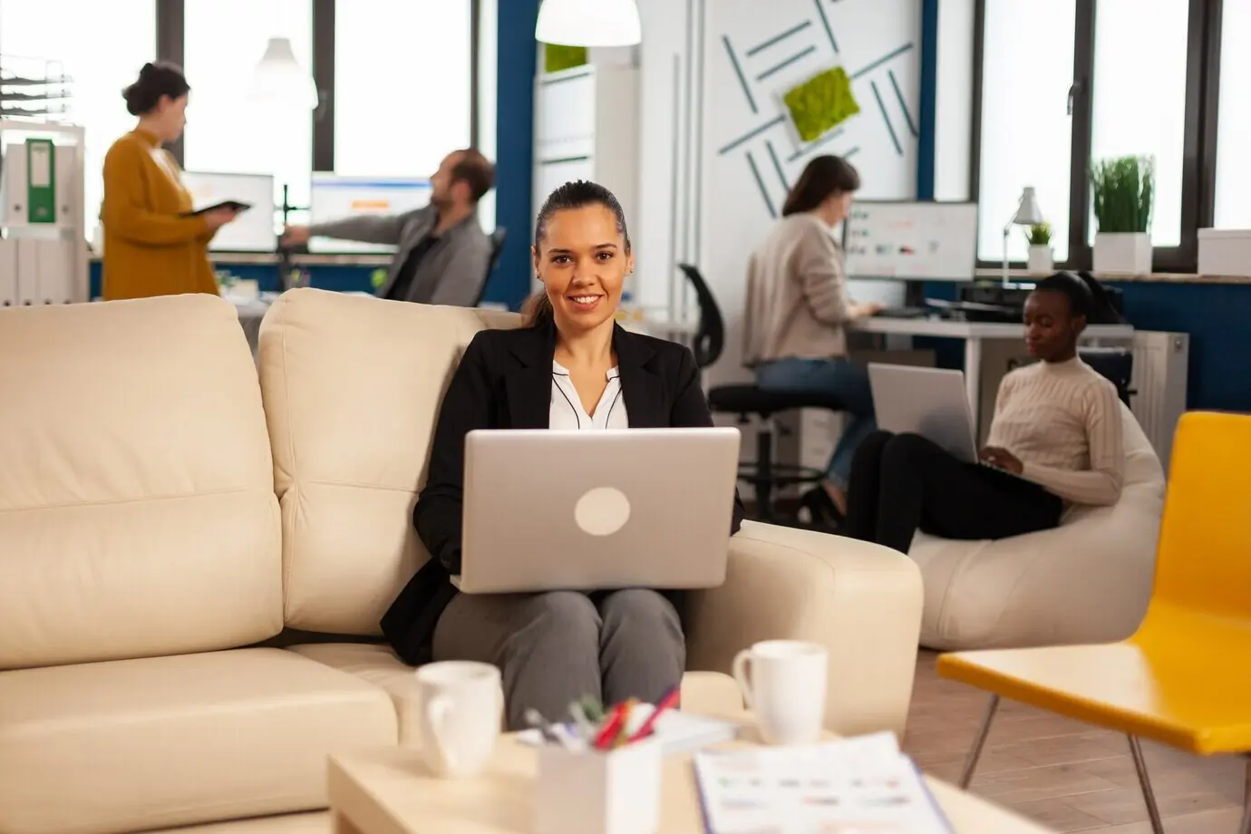 Female manager typing on a laptop, smiling at the camera, with diverse colleagues working in the background.