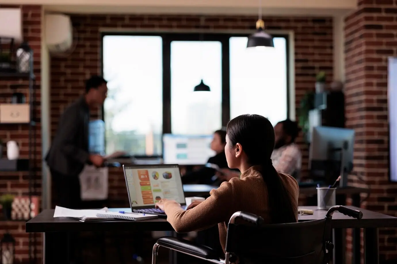 An Asian businesswoman living with a disability analyzes a report on a laptop, working with statistics in a disability-friendly office. She is an employee suffering from a chronic impairment and uses a wheelchair.
