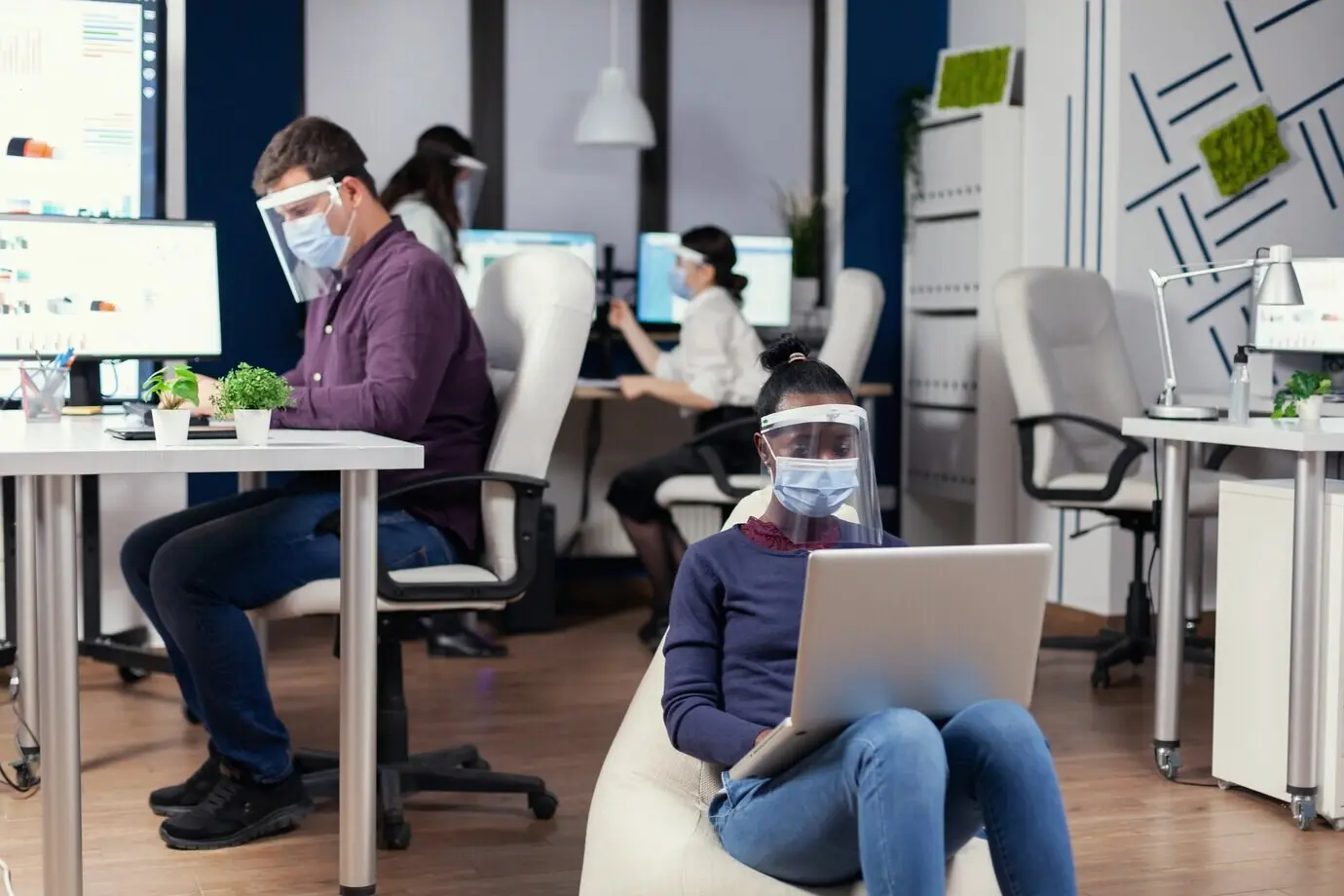 Black freelancer wearing a protective mask against coronavirus sits in an armchair in the middle of an office room, analyzing a project on a digital tablet. A multiethnic business team works while respecting social distance.